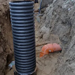 Głęboki wykop z widocznymi czarnymi rurami karbowanymi pionowo oraz pomarańczowymi rurami kanalizacyjnymi w piasku, łopata wbita w ziemię, budynek w tle.