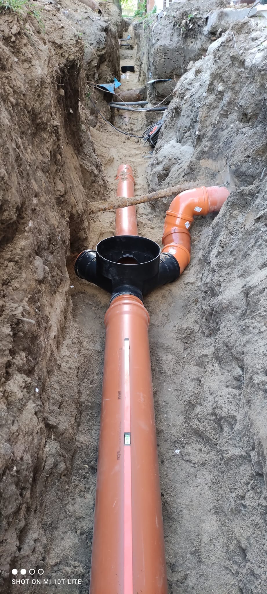 Pomarańczowe rury kanalizacyjne z czarnym trójnikiem w wykopie ziemnym, widoczny poziomica na rurze.