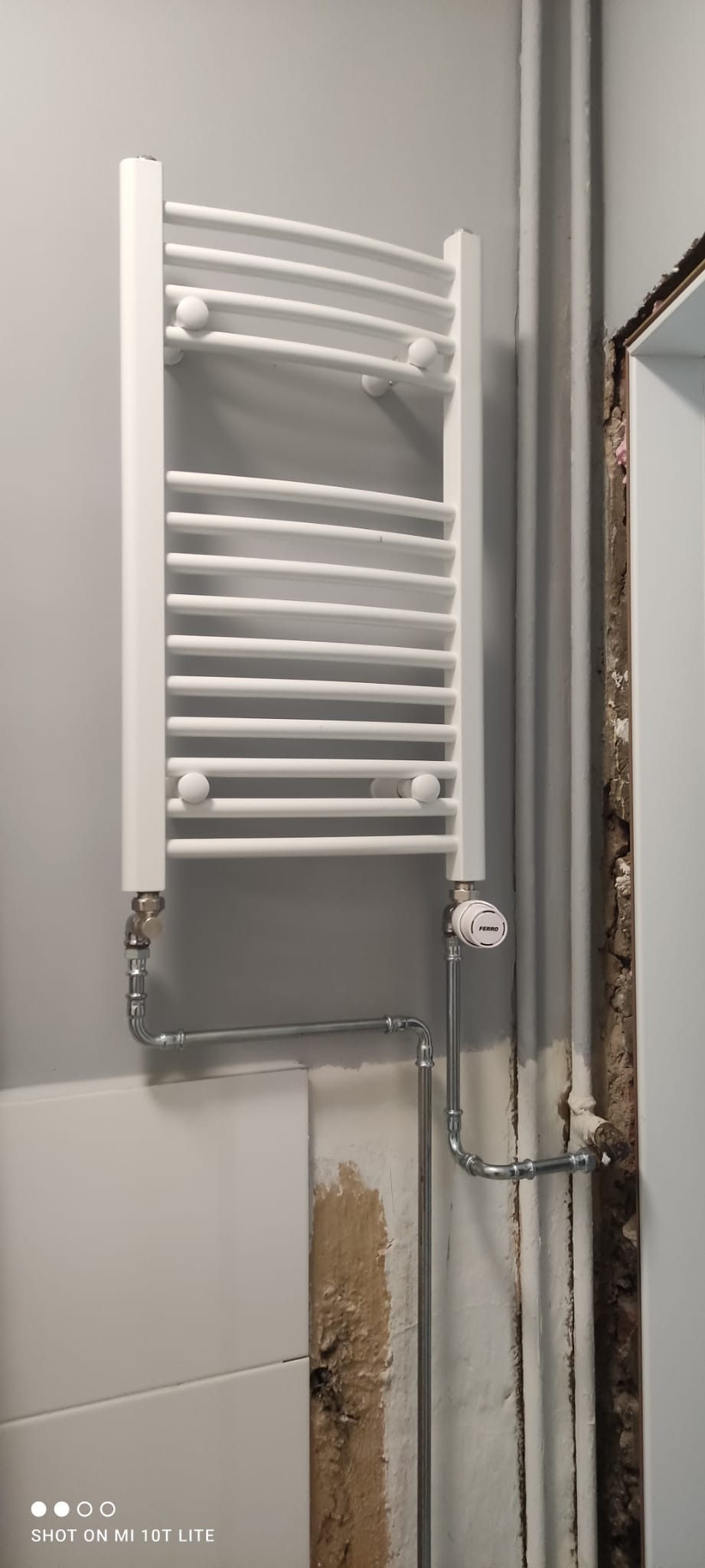 Biały, drabinkowy grzejnik łazienkowy z termostatem Ferro, zamontowany na szarej ścianie, z widocznymi rurami instalacji grzewczej i fragmentem obudowy drzwi z ubytkami tynku.