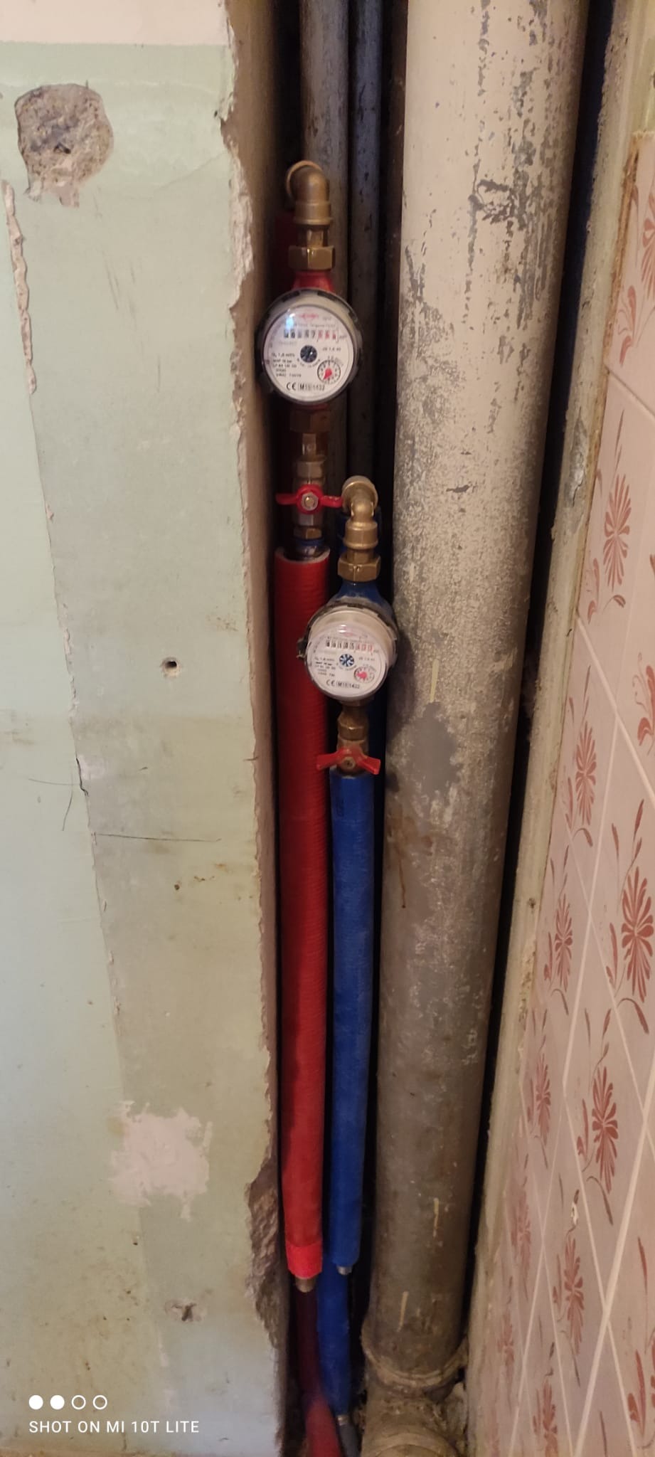 Dwa liczniki wody z czerwonym i niebieskim ociepleniem na rurach, zamontowane w wąskiej przestrzeni przy ścianie z odpadającym tynkiem i starymi płytkami.