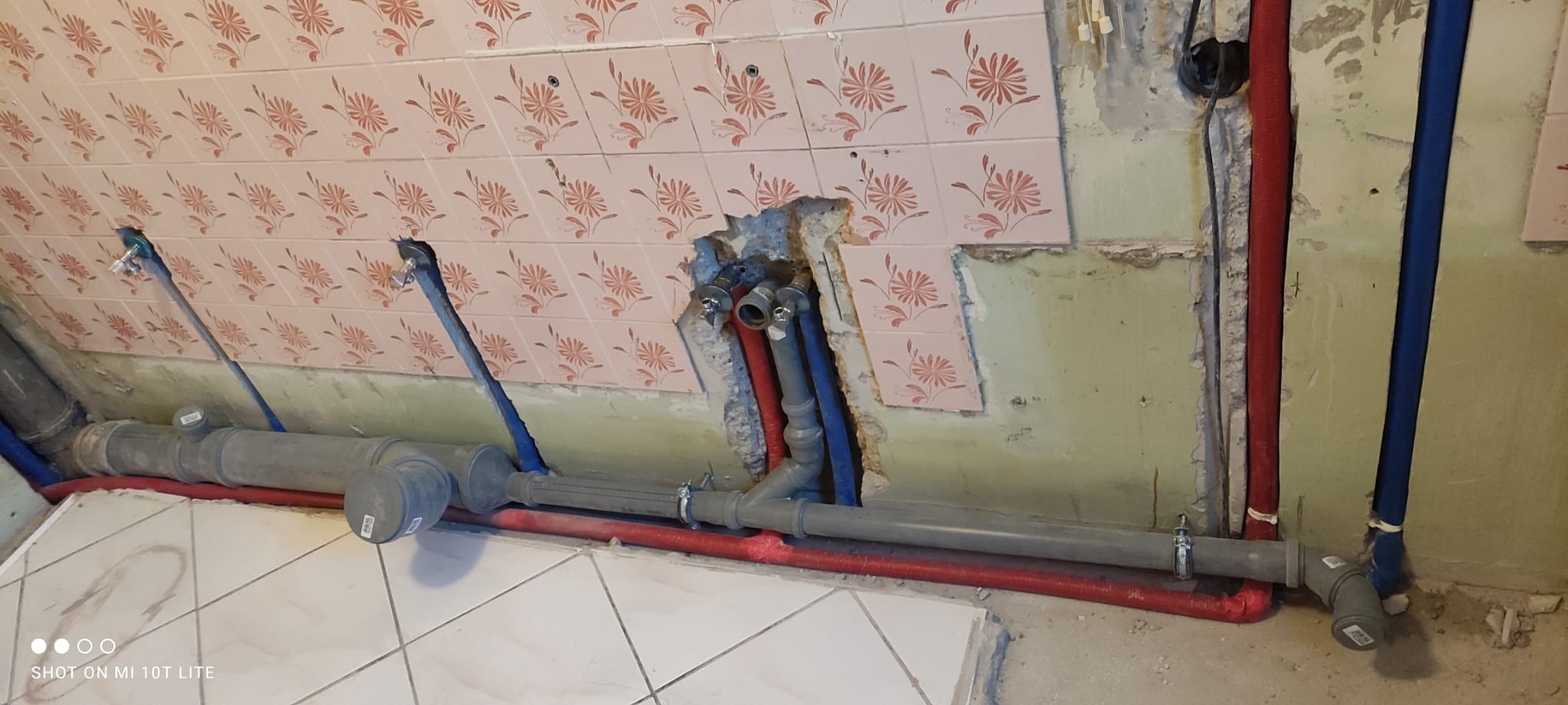 Niedokończone przyłącze wodno-kanalizacyjne z widocznymi rurami, fragmentami płytek i surową ścianą.