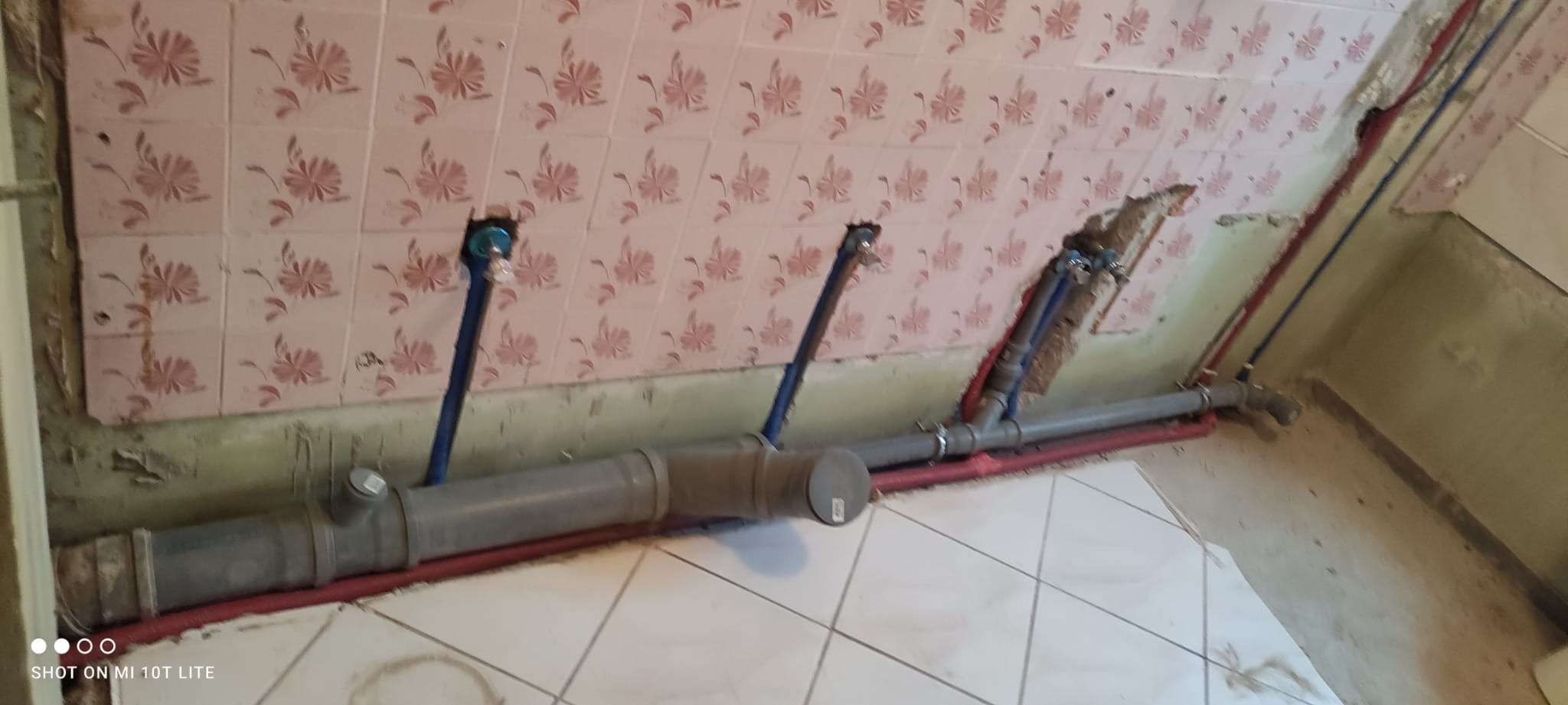 Szare rury kanalizacyjne i niebieskie rury wodne zamontowane przy ścianie z różowymi płytkami w trakcie remontu łazienki.