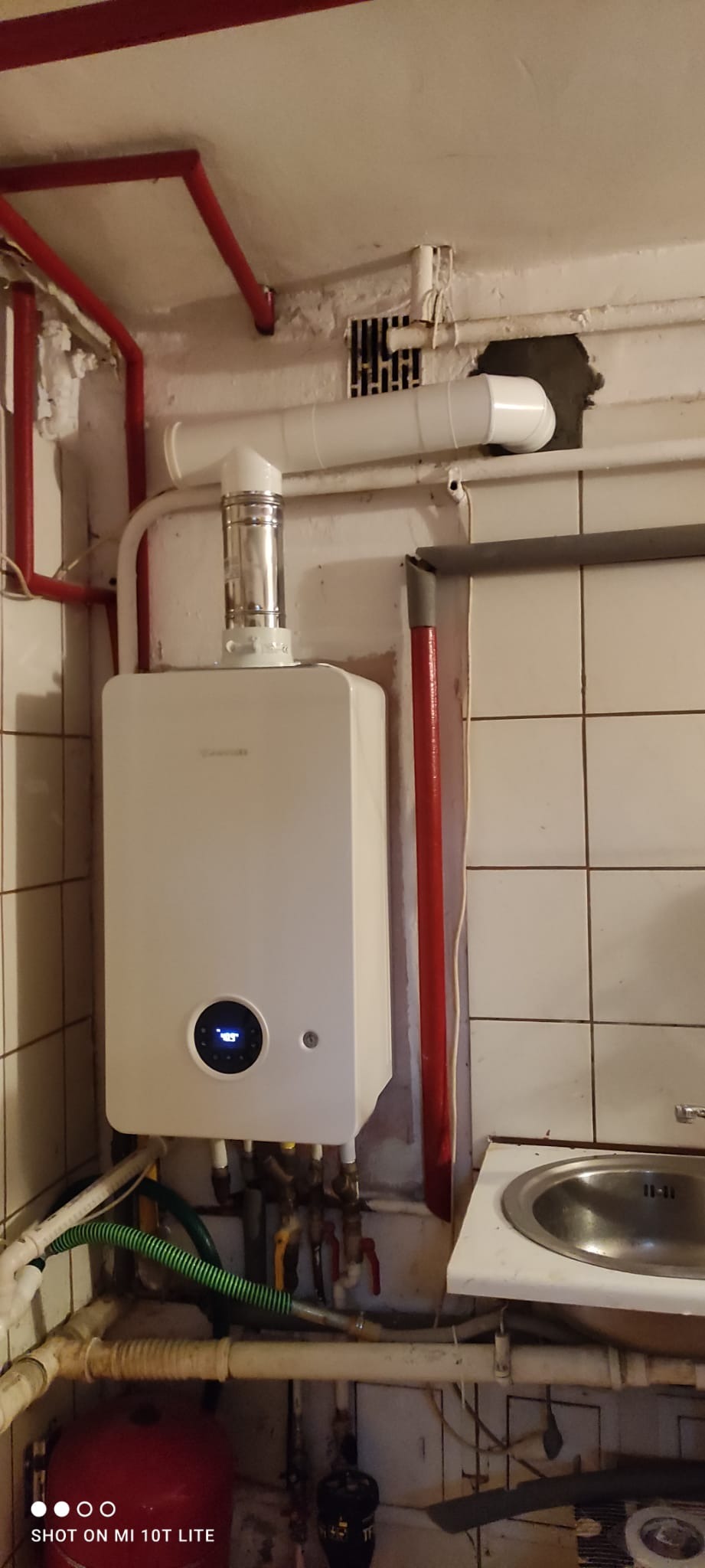 Biały piec gazowy z podłączonymi rurami i kominem, zamontowany na ścianie w pomieszczeniu z płytkami, widoczne rury centralnego ogrzewania i umywalka.