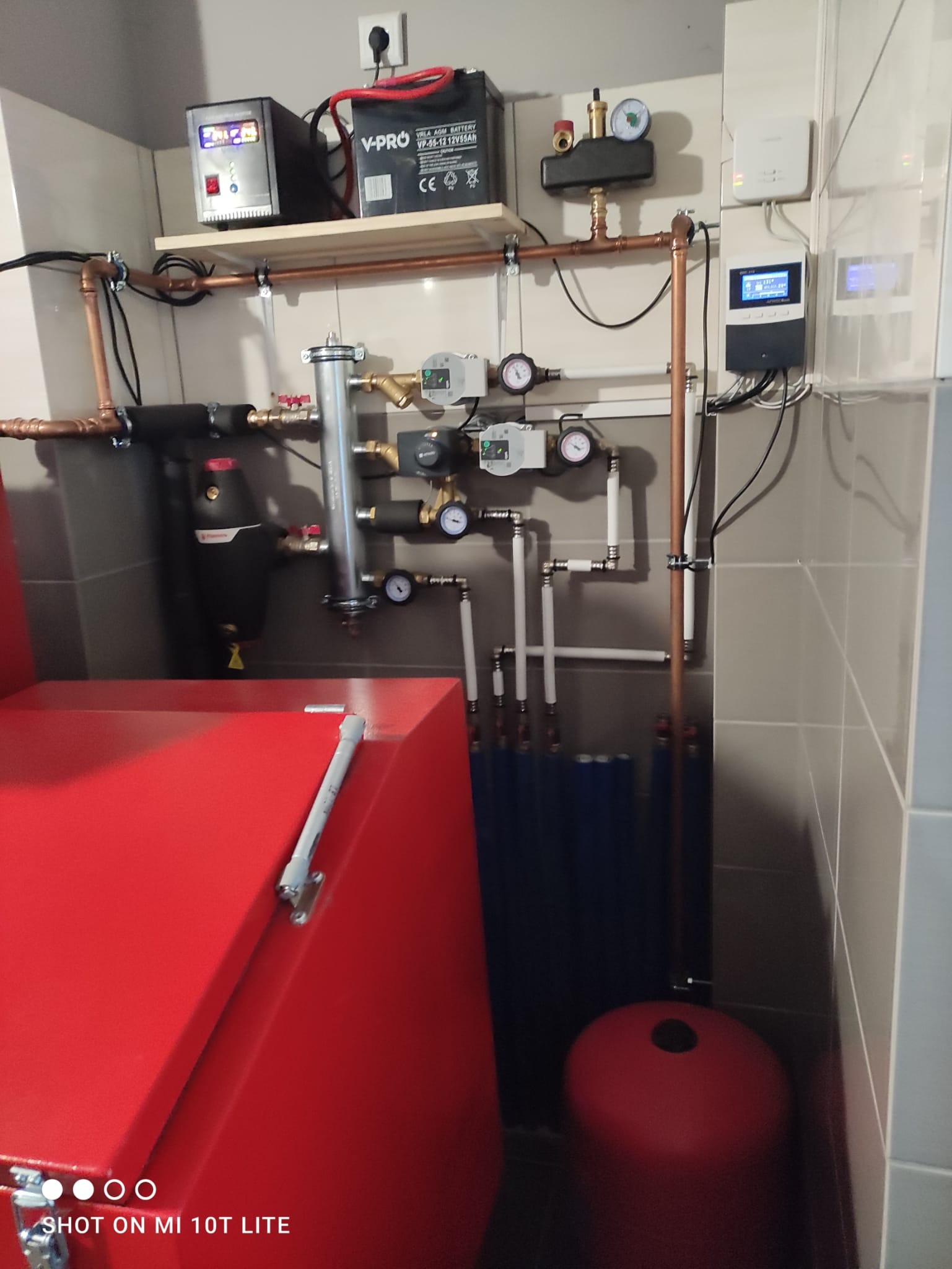 Czerwony piec CO z podłączoną instalacją hydrauliczną, widoczne rury miedziane, zawory, manometry, zbiornik wyrównawczy i sterownik na ścianie z szarych płytek.