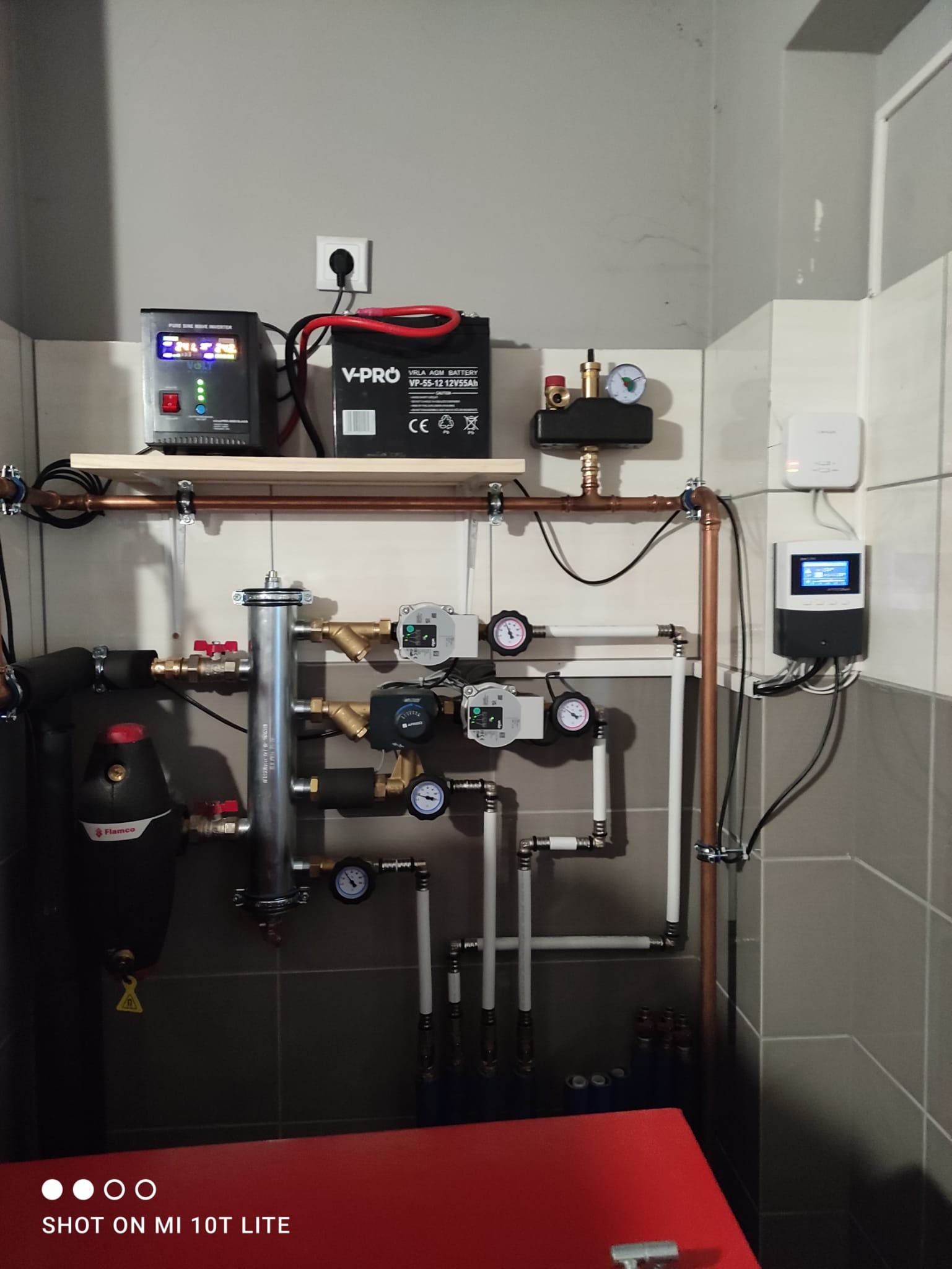 Złożony system instalacji hydraulicznej z zbiornikiem Flamco, pompami obiegowymi, manometrami, rurami miedzianymi i plastikowymi, zasilaczem awaryjnym i sterownikiem na ścianie z szarymi płytkami.