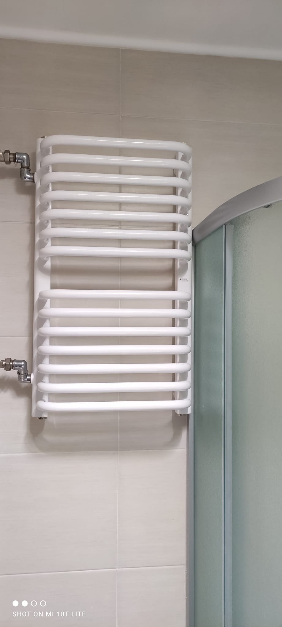 Biały, drabinkowy grzejnik łazienkowy zamontowany na jasnej, kafelkowanej ścianie, obok kabiny prysznicowej z mleczną szybą.