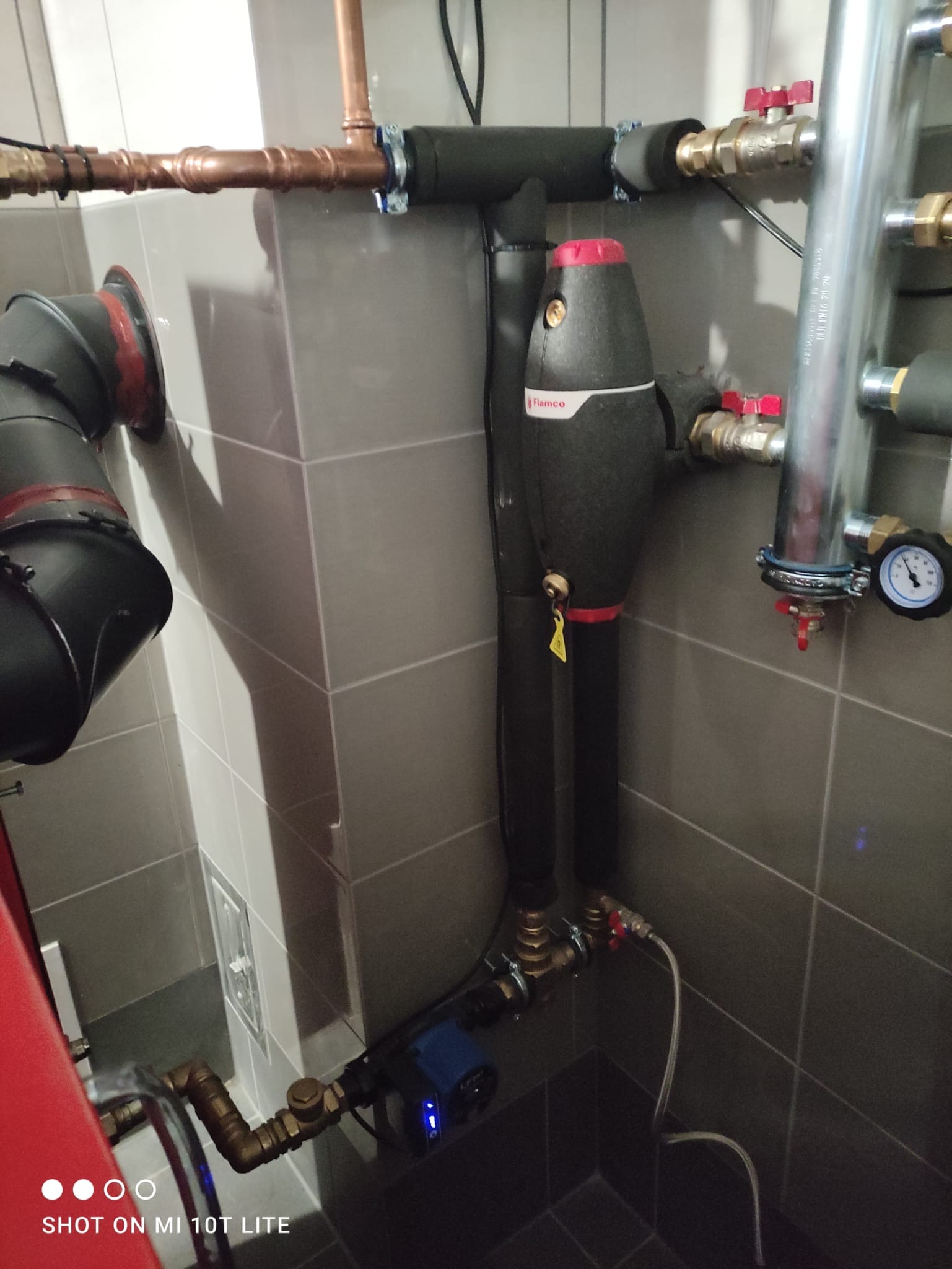 Szare płytki łazienkowe, instalacja hydrauliczna z rurami miedzianymi, czarnymi izolacjami i zaworami z czerwonymi pokrętłami, widoczny manometr i urządzenie Flämco z żółtą zawieszką.