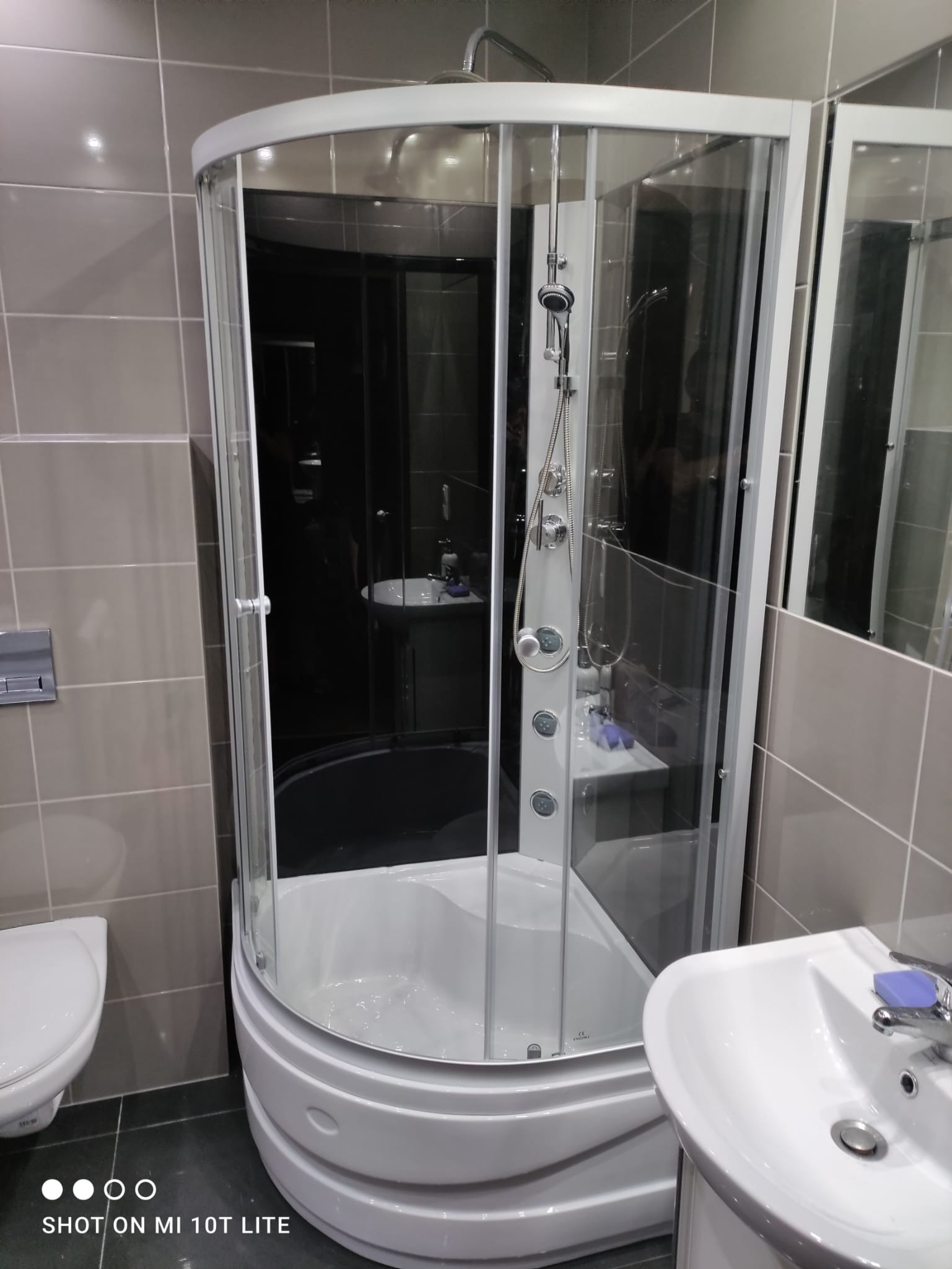 Nowoczesna kabina prysznicowa z hydromasażem, widoczna umywalka z mydłem i toaleta w łazience wyłożonej szarymi płytkami.