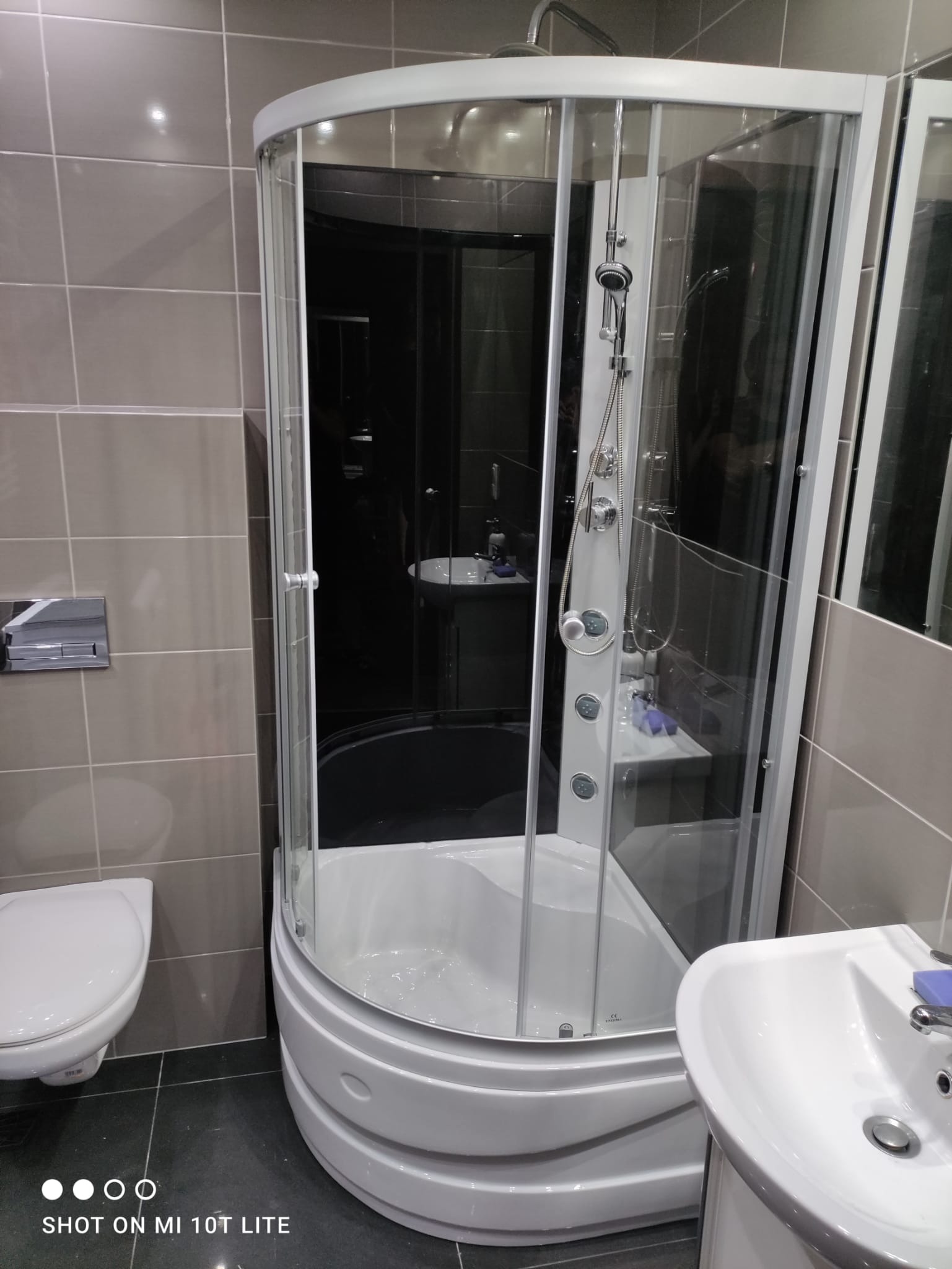 Kabina prysznicowa z hydromasażem, umywalka i toaleta w łazience wykończonej szarymi płytkami.