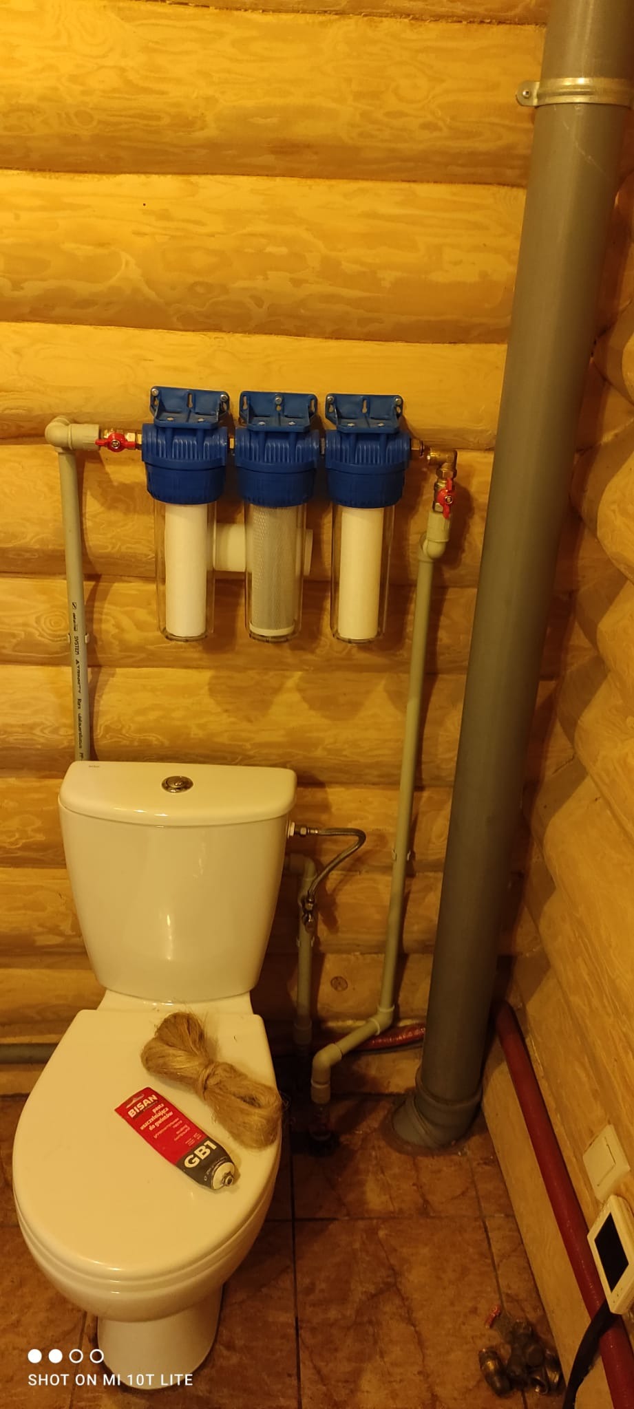 Wnętrze łazienki wykończonej drewnem, z zamontowanym filtrem wody z trzema przezroczystymi obudowami i niebieskimi głowicami, wiszącym nad toaletą, na której leży pakuła i uszczelniacz w tubce.