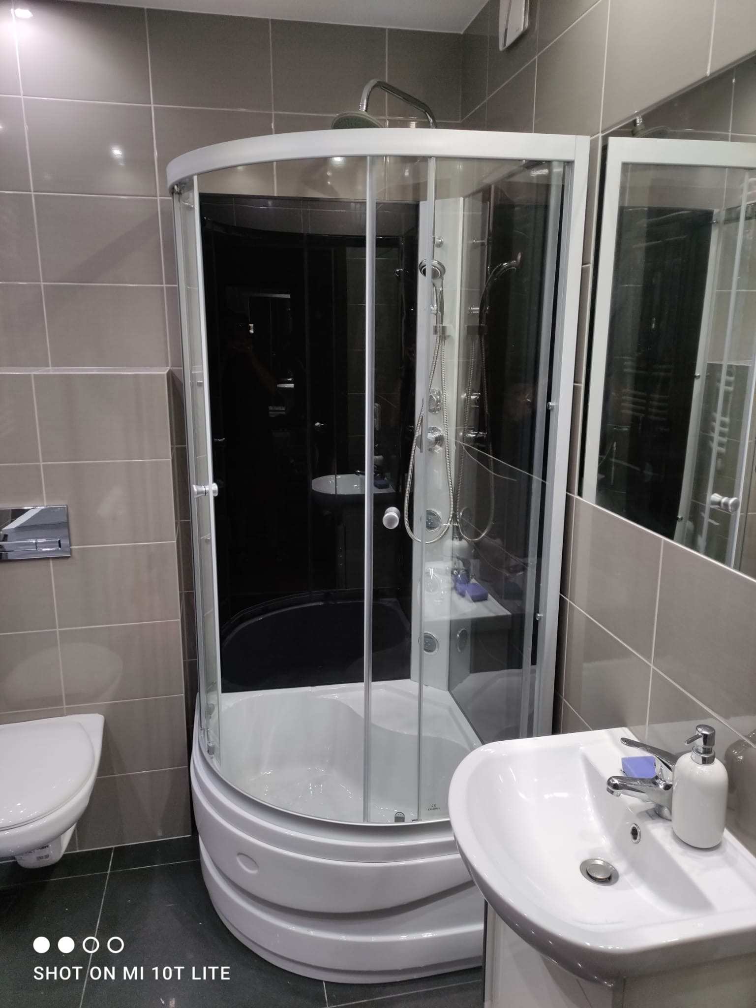 Nowoczesna kabina prysznicowa narożna z ciemnym szkłem, umywalka z mydłem i dozownikiem, szare płytki w łazience.