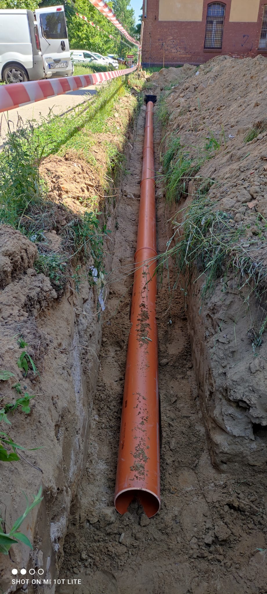 Pomarańczowa rura kanalizacyjna ułożona w wykopie ziemnym, zabezpieczonym taśmą ostrzegawczą, widok z góry, Olsztyn.