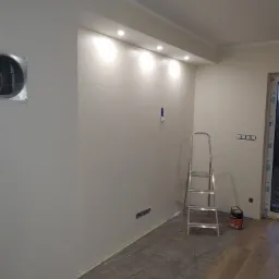 Wnętrze w trakcie prac w Koszalinie: ściany przygotowane do malowania, zabezpieczona podłoga, drabina i puszka farby w rogu pomieszczenia.