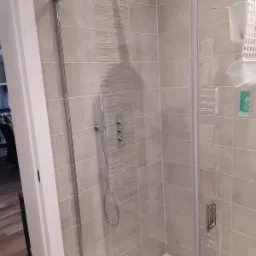 Kabina prysznicowa z deszczownicą i szklanymi drzwiami, wykończona płytkami w odcieniach szarości z dekoracyjnym pasem, widoczny zestaw prysznicowy z termostatem i półka na kosmetyki.