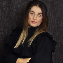 Portret kobiety z długimi, brązowo-blond włosami, ubrana w czarną bluzkę z falbanami, na ciemnoszarym teksturowanym tle.