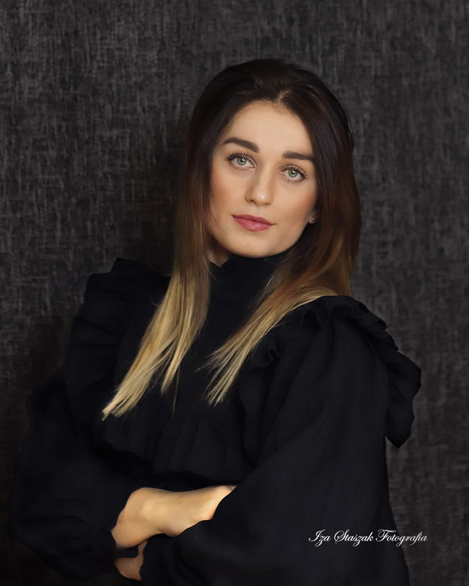 Portret kobiety z długimi, brązowo-blond włosami, ubrana w czarną bluzkę z falbanami, na ciemnoszarym teksturowanym tle.