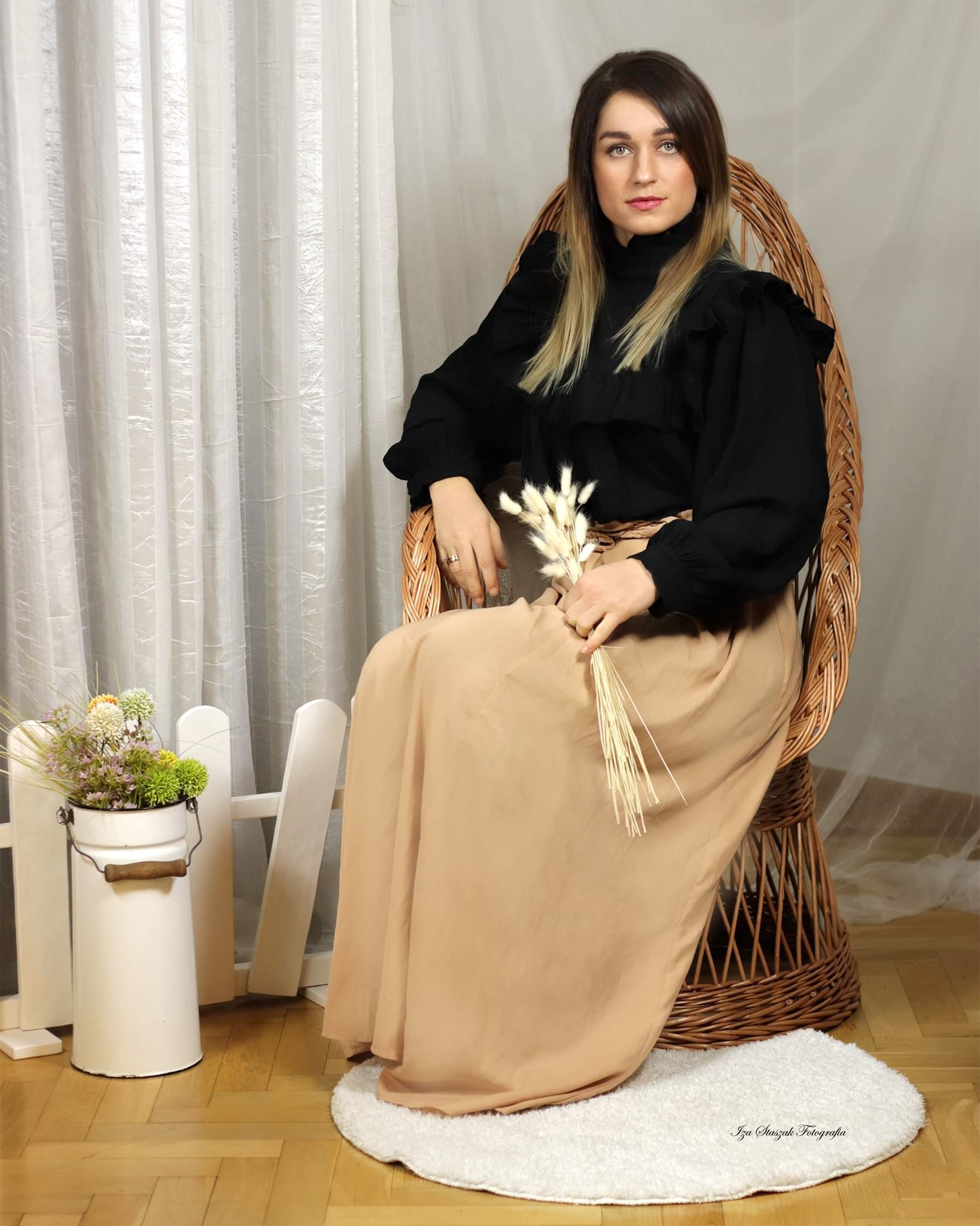 Kobieta w czarnej bluzce i beżowej spódnicy siedzi w wiklinowym fotelu, trzymając w dłoniach bukiet suszonych traw, obok dekoracja z białym płotkiem i kwiatami w emaliowanym wiaderku, w tle zwiewna...