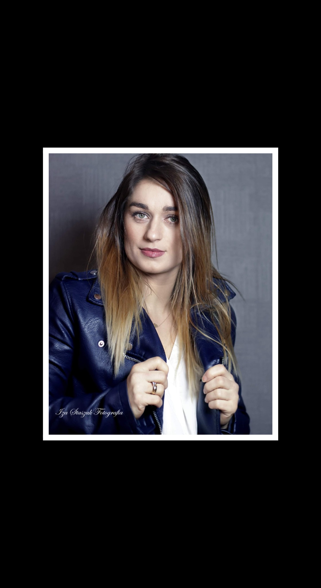 Portret kobiety w niebieskiej skórzanej kurtce, z delikatnym makijażem i pierścionkiem na palcu, na szarym tle.