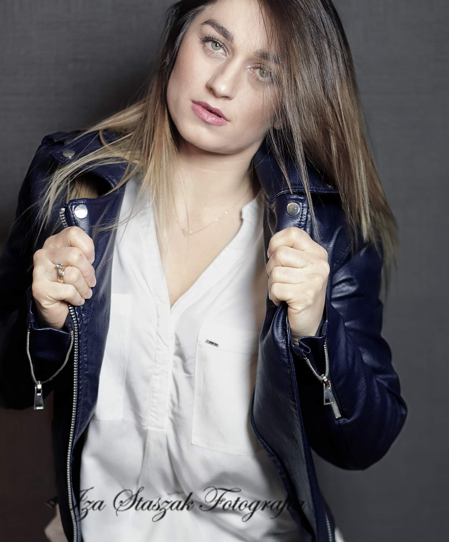 Portret kobiety w białej koszuli i granatowej skórzanej kurtce, z pierścionkiem na palcu, na szarym tle. Widoczne logo fotografa w dolnej części kadru.