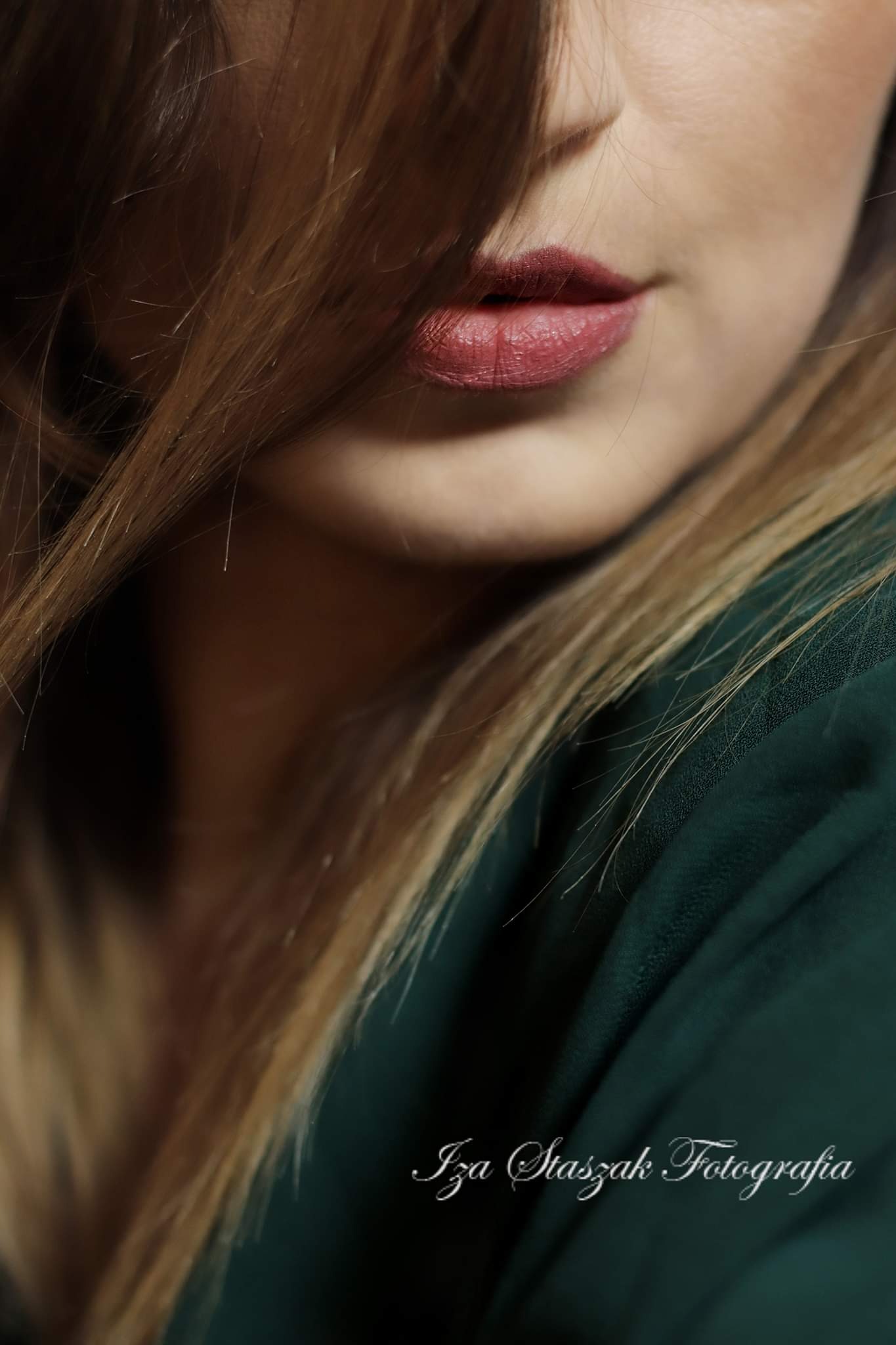 Zbliżenie na usta kobiety z rozpuszczonymi włosami, w tle ciemnozielona tkanina i widoczne logo fotografa.