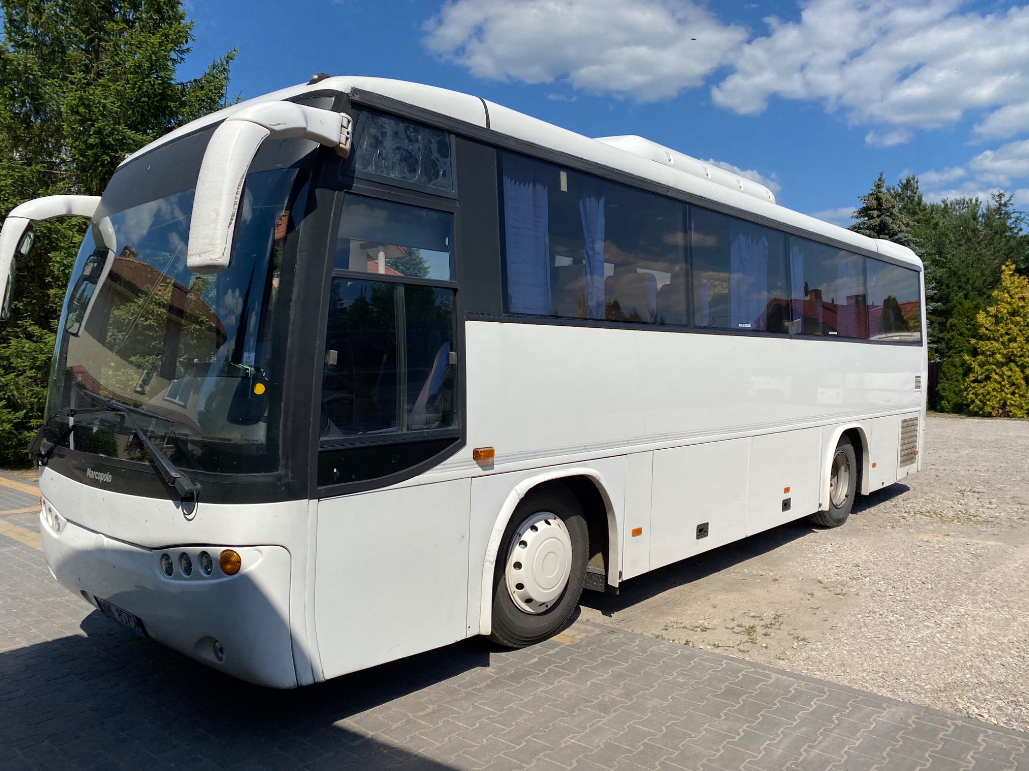 Biały autokar marki Marcopolo zaparkowany na placu z kostki brukowej, widok z przodu i boku, z widocznymi zasłonami w oknach.