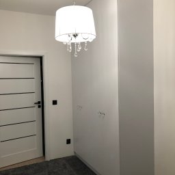 Szafa pod wymiar - Wysoka, biała szafa wnękowa z kryształowymi gałkami w pokoju z szarym dywanem i drzwiami z czarnymi akcentami. Nad szafą wisi ozdobny żyrandol.