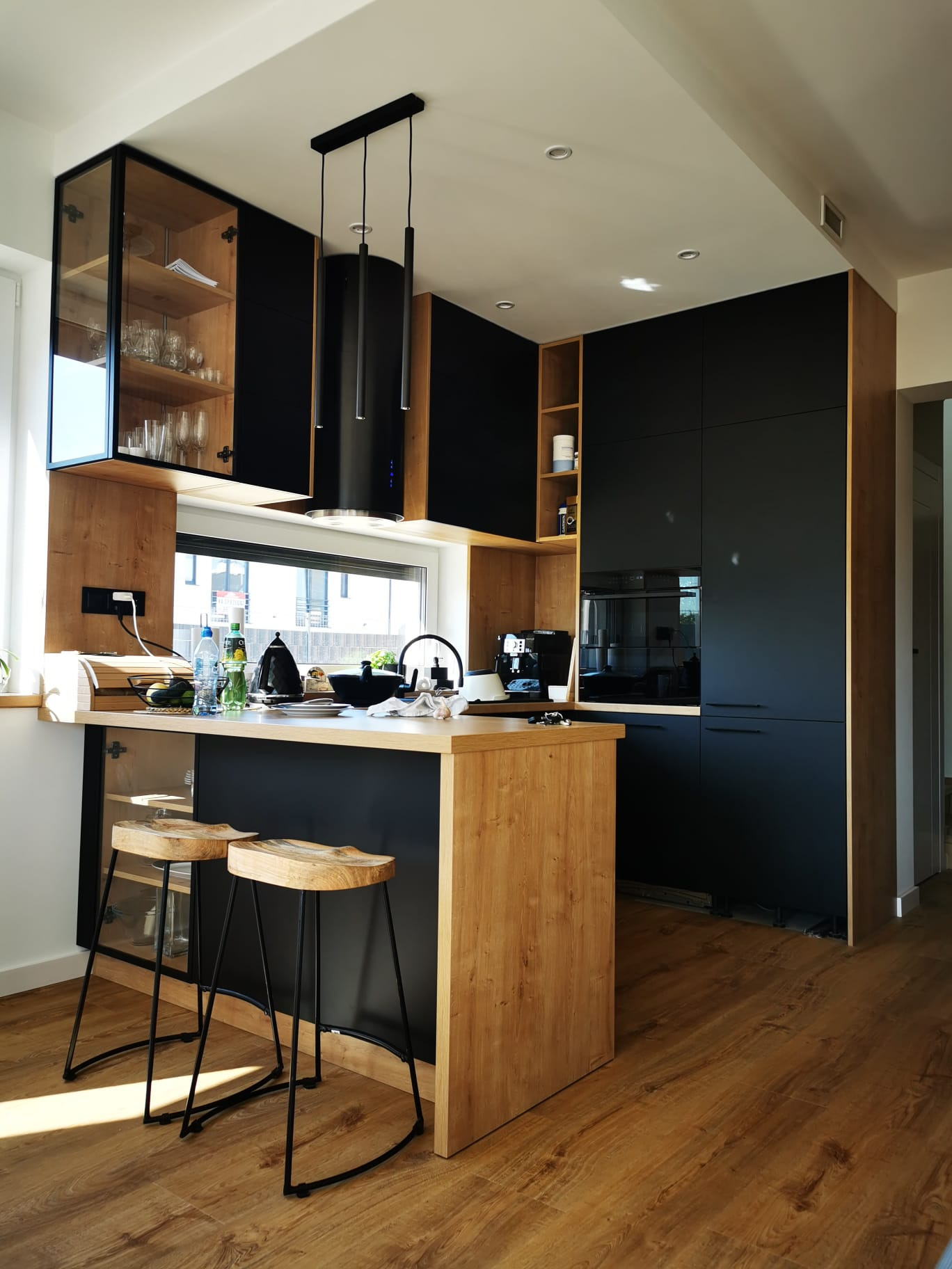 Nowoczesna kuchnia z czarnymi frontami i drewnianymi akcentami, wyspa z dwoma stołkami barowymi, szklane witryny górnych szafek, panoramiczne okno.