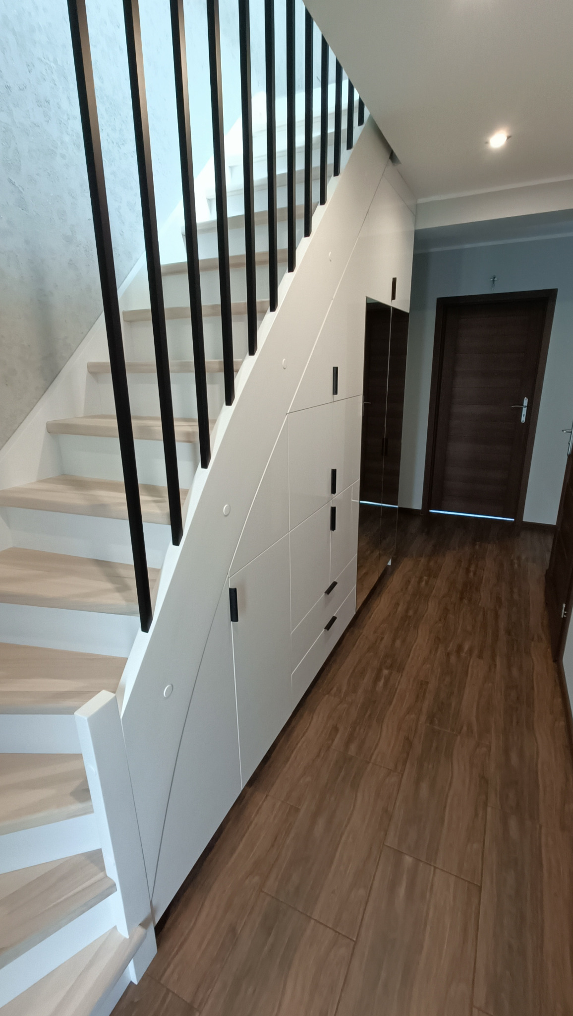Biała szafa wnękowa pod schodami z czarnymi uchwytami, lustrzanym panelem i drewnianymi schodami z czarną balustradą w jasnym wnętrzu.