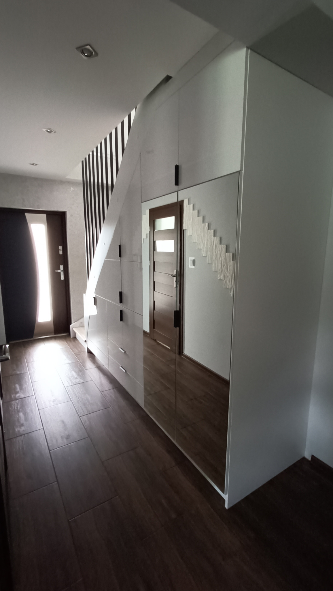 Biała szafa wnękowa pod schodami z lustrzanymi drzwiami, odbijająca wnętrze domu. Ciemna podłoga z płytek, widoczny fragment schodów z czarnymi barierkami.