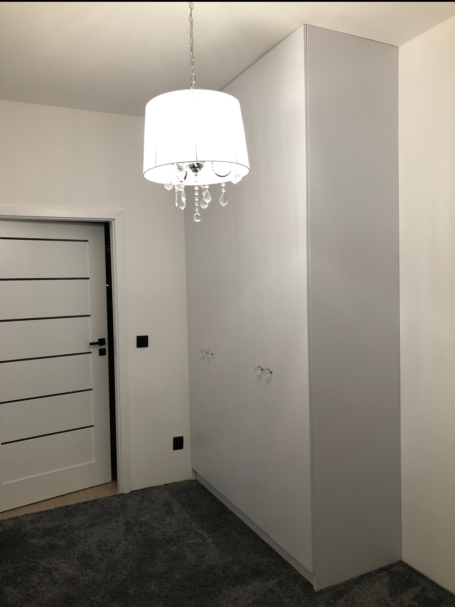 Wysoka, biała szafa wnękowa z kryształowymi gałkami w pokoju z szarym dywanem i drzwiami z czarnymi akcentami. Nad szafą wisi ozdobny żyrandol.