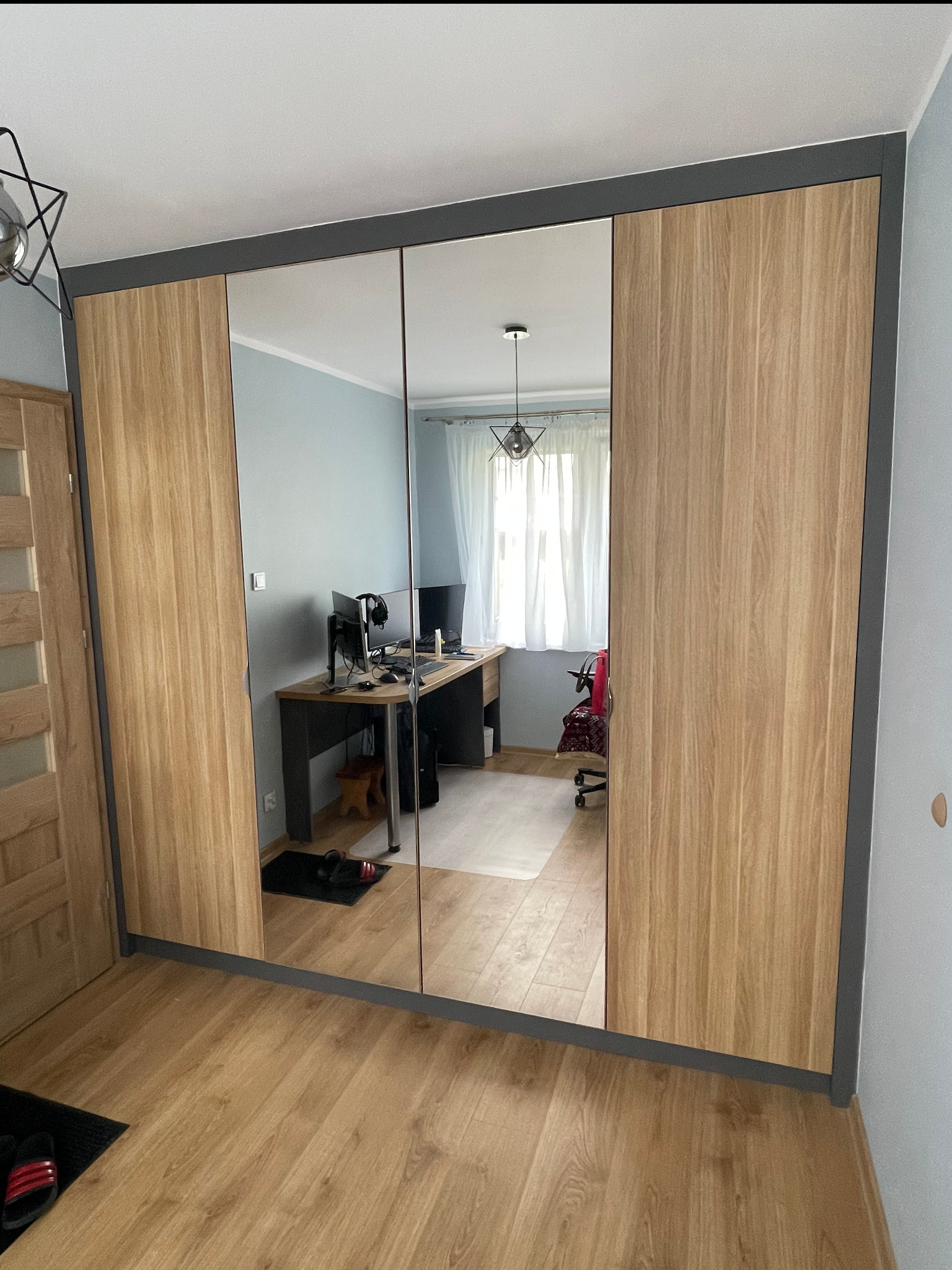 Trzydrzwiowa szafa wnękowa z lustrami i drewnianymi bokami, szare obramowanie, widok na biurko z komputerem odbijające się w lustrach.