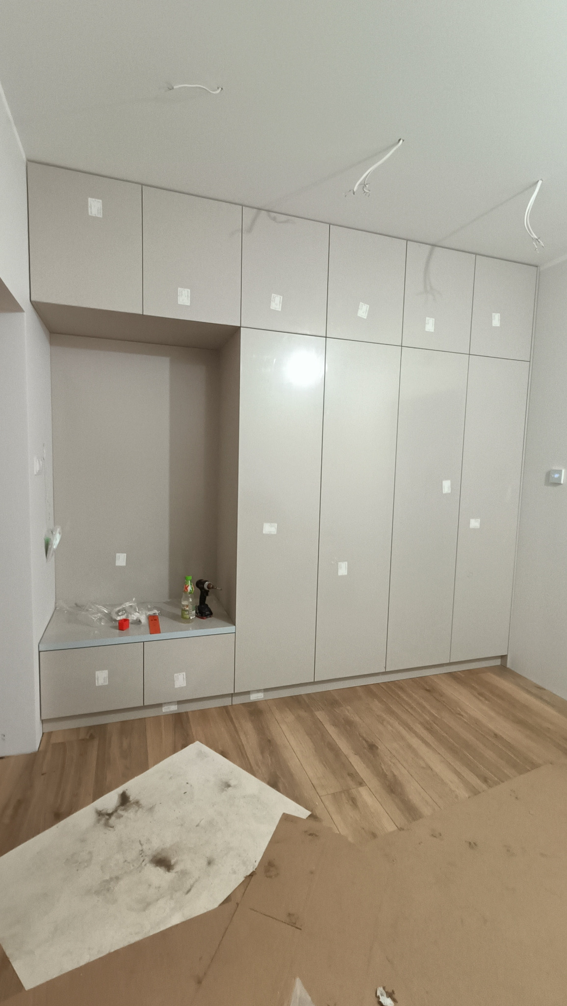 Jasna szafa wnękowa z otwartą półką i błyszczącymi frontami, zamontowana w pokoju z drewnianą podłogą. Na półce widoczne narzędzia i akcesoria montażowe.