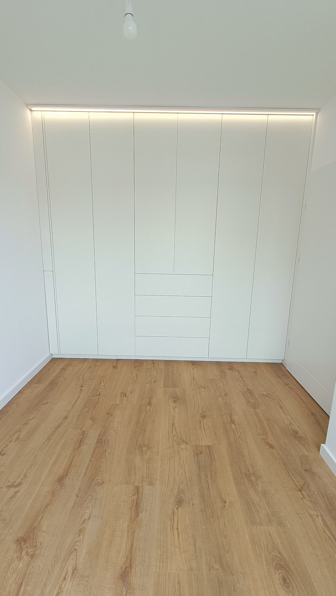 Biała szafa wnękowa z oświetleniem LED w górnej części, widok na drewnianą podłogę. Minimalistyczny design, nowoczesne wykończenie.