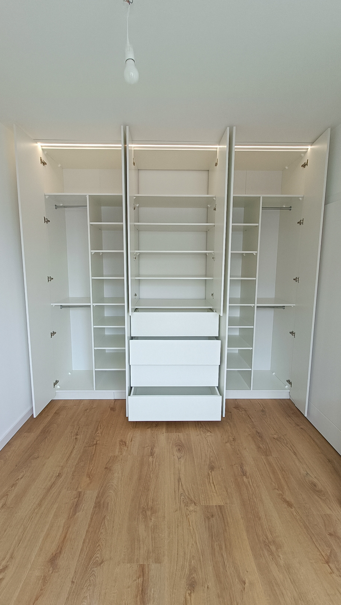 Biała szafa wnękowa z otwartymi drzwiami i szufladami, oświetlenie LED u góry, podłoga z paneli. Widok z przodu, wnętrze szafy z półkami i drążkami.