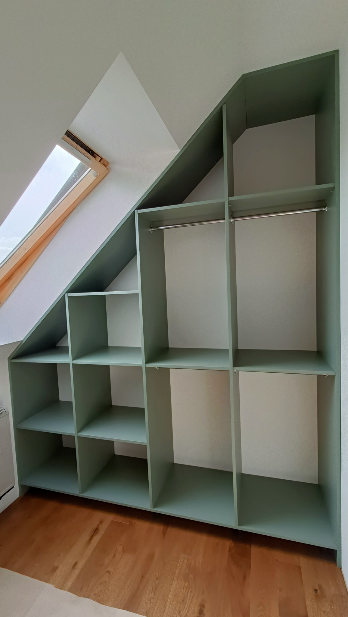 Szafa wnękowa z półkami i drążkami, dopasowana do skosu dachu, w odcieniach zieleni, na podłodze z paneli drewnianych, z oknem dachowym w tle.