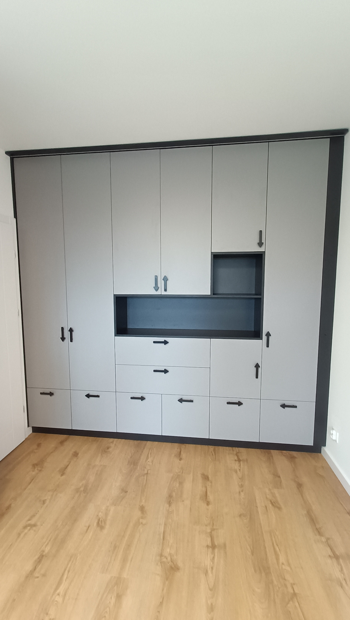 Szara szafa wnękowa z czarnymi akcentami i nietypowymi uchwytami w kształcie strzałek, na tle drewnianej podłogi i białych ścian. Minimalistyczny design.
