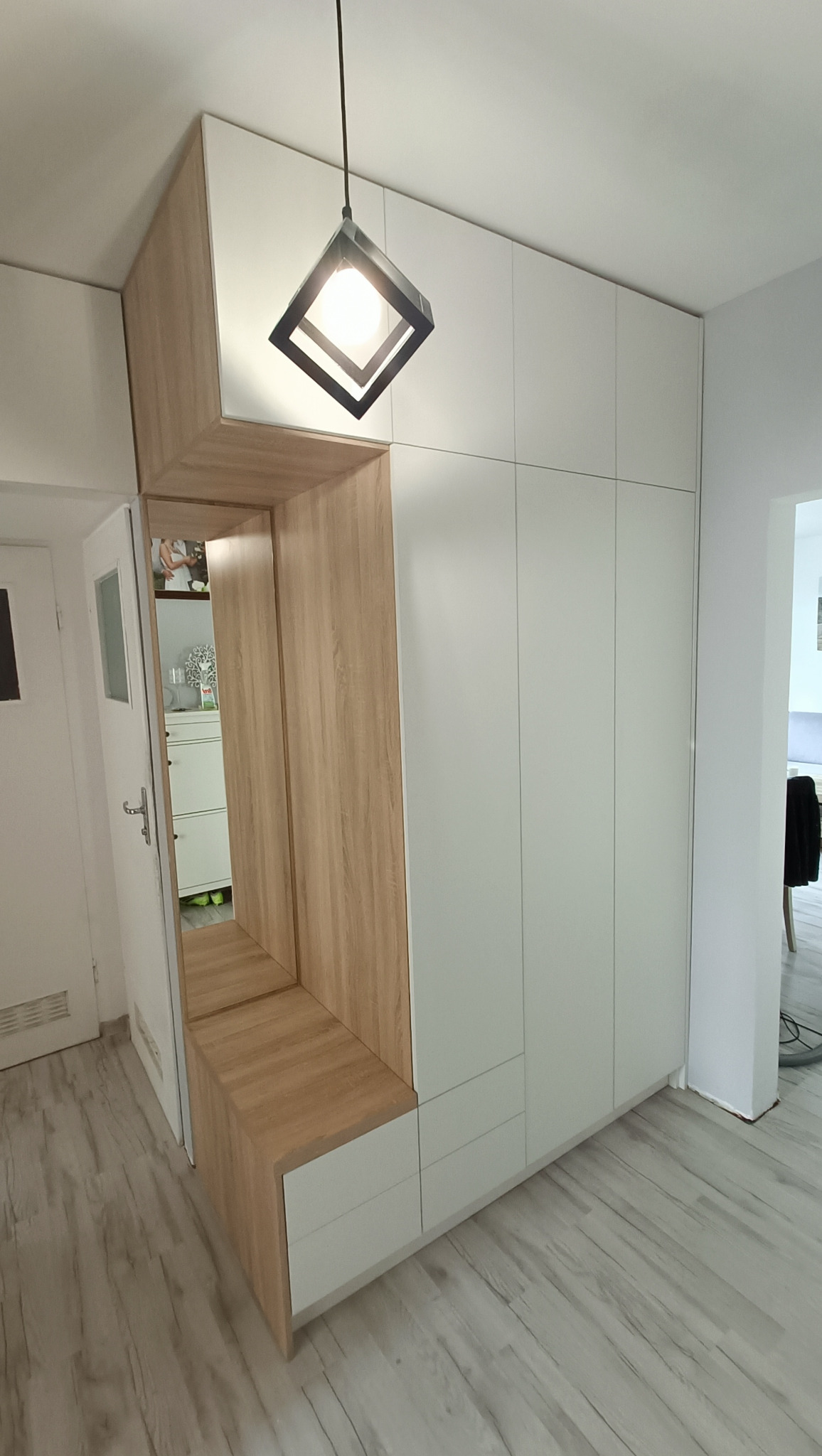 Biała szafa wnękowa z elementami drewnopodobnymi, siedziskiem i lustrem w przedpokoju. Nowoczesna lampa sufitowa. Jasne panele podłogowe.