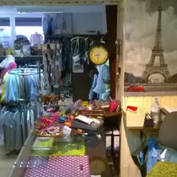 Wnętrze małego sklepu odzieżowego z różnorodną odzieżą na wieszakach, biżuterią na ladzie i plakatem z wieżą Eiffla w tle.