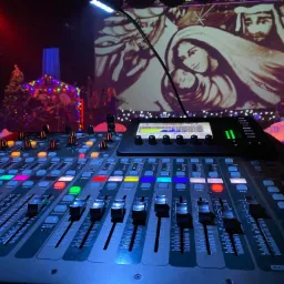 Profesjonalny mikser audio na pierwszym planie, z podświetlanymi przyciskami i suwakami, w tle scena z projekcją przedstawiającą scenę biblijną, oświetlona dekoracyjnymi lampkami i choinką...