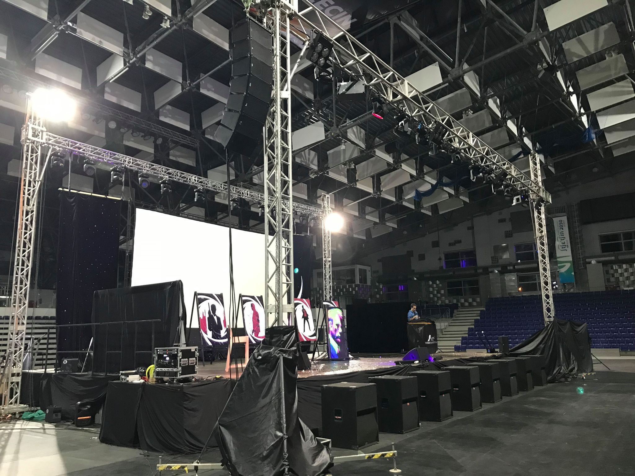 Scena z podwieszonym systemem nagłośnieniowym line array, oświetleniem punktowym i ekranem projekcyjnym, przygotowana do wydarzenia w hali widowiskowej.