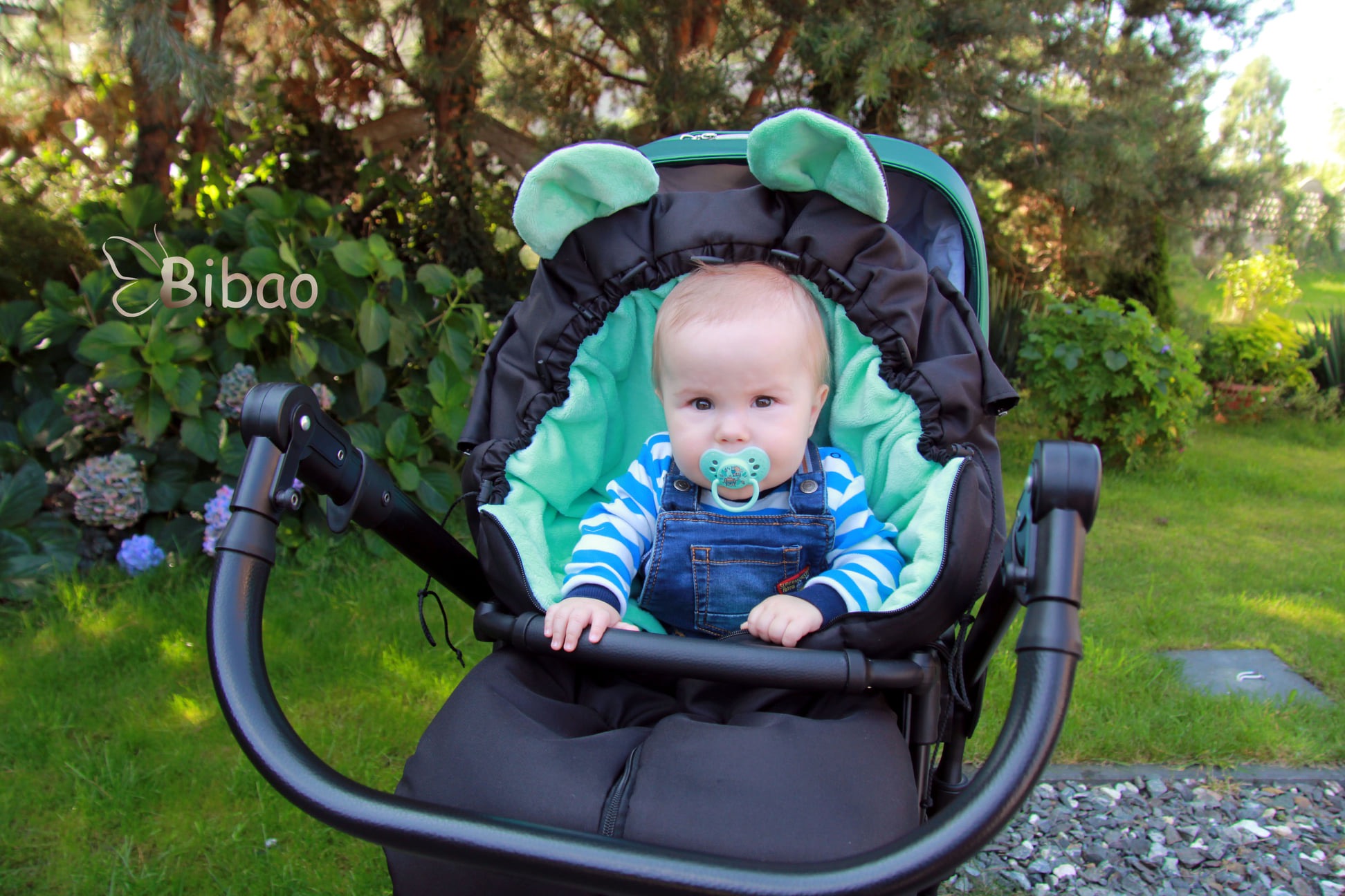 Dziecko w wózku z czarnym śpiworkiem i miętową wyściółką z uszami misia, na tle zielonego ogrodu.