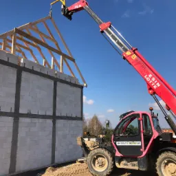 Czerwona koparka teleskopowa z żółtym osprzętem podnosi drewnianą konstrukcję dachu na budynek z białych bloczków, widoczne numery kontaktowe na kabinie.