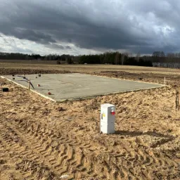 Wylana płyta fundamentowa na gruncie, widoczne wystające rury i elementy instalacji, w tle pole i pochmurne niebo.