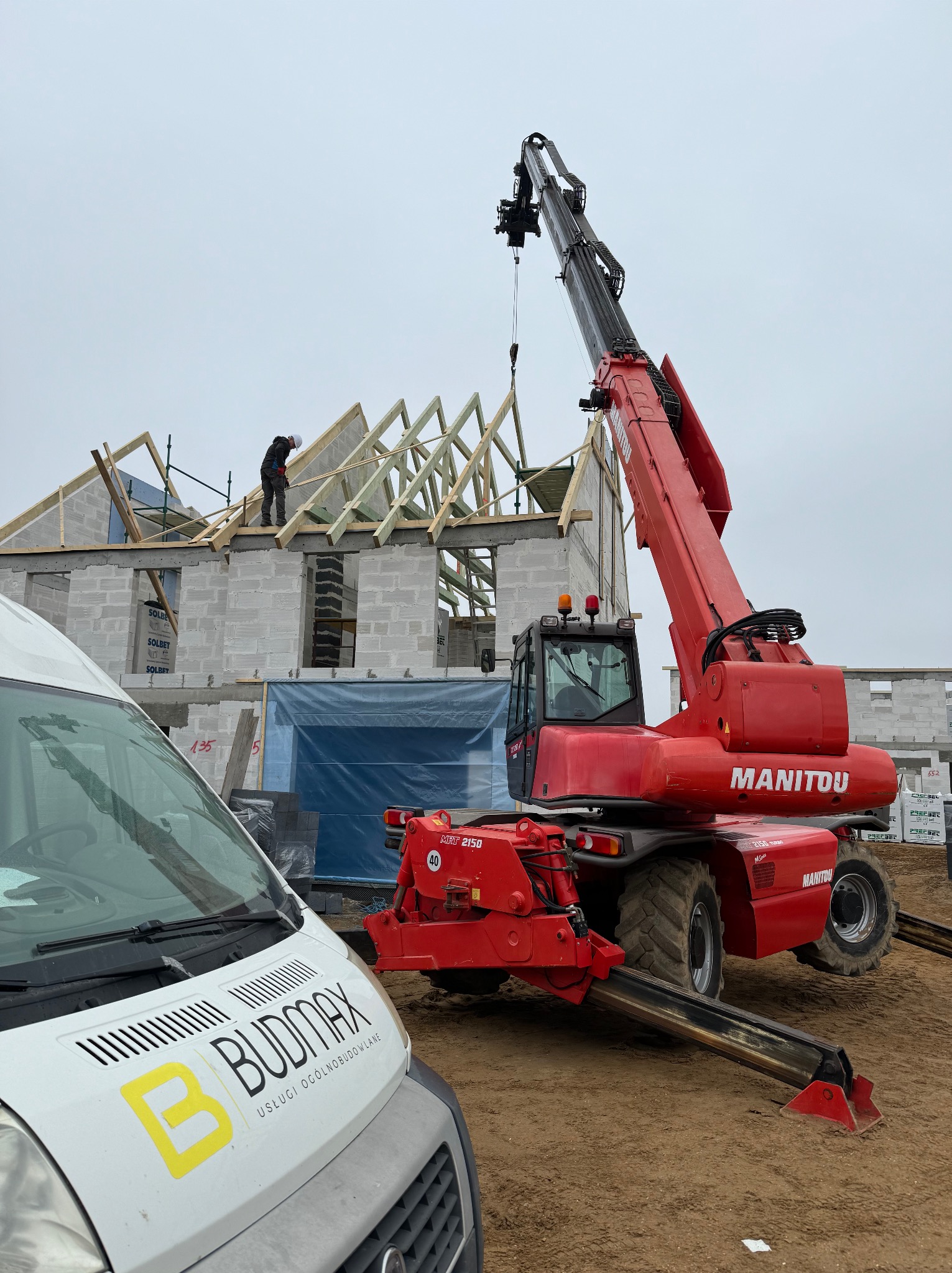 Czerwona koparka teleskopowa Manitou podnosi element drewniany na budowę domu z białego bloczka, w tle pracownik w kasku na dachu, z przodu samochód dostawczy z logo firmy Budmax.