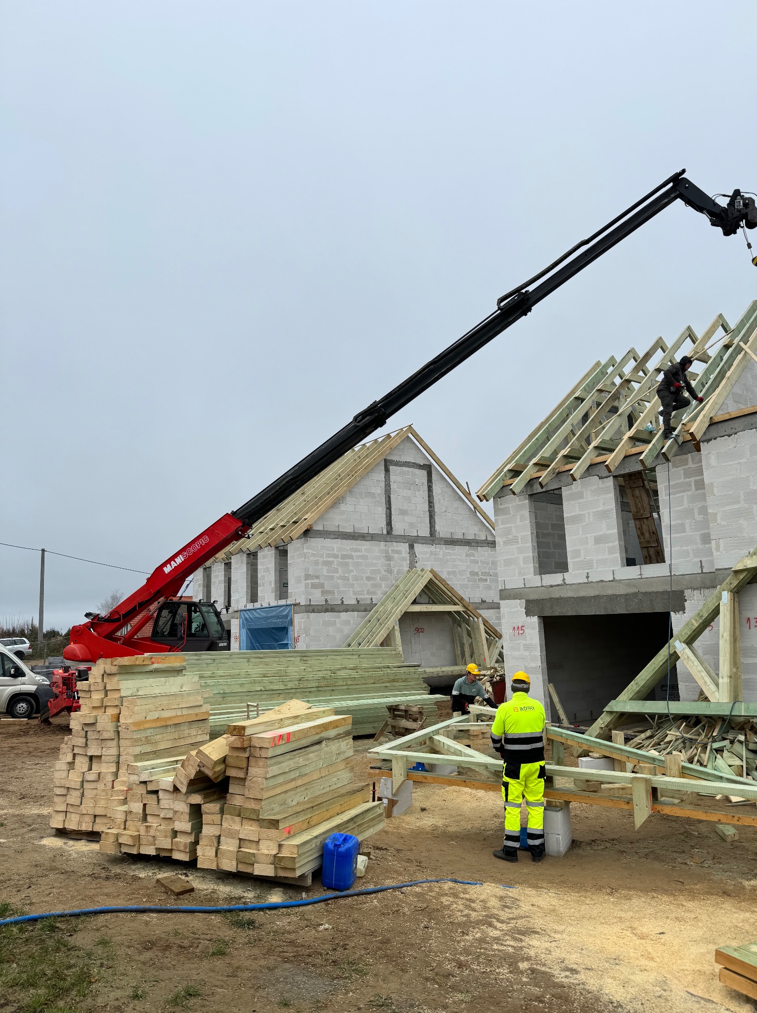 Wznoszenie konstrukcji dachowej na dwóch budynkach z bloczków betonowych, widoczny żuraw teleskopowy i pracownicy w odzieży roboczej.