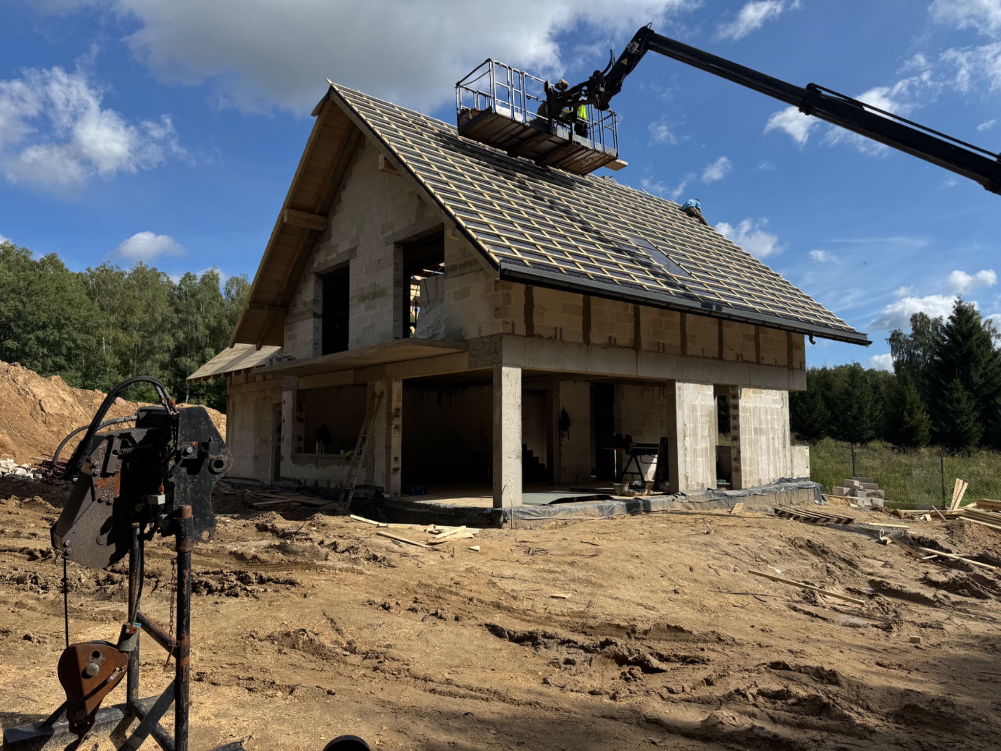 Budowa domu z bloczków: surowy stan z drewnianą konstrukcją dachu i dźwigiem. Widoczne niezabudowane otwory okienne i drzwiowe, teren budowy.
