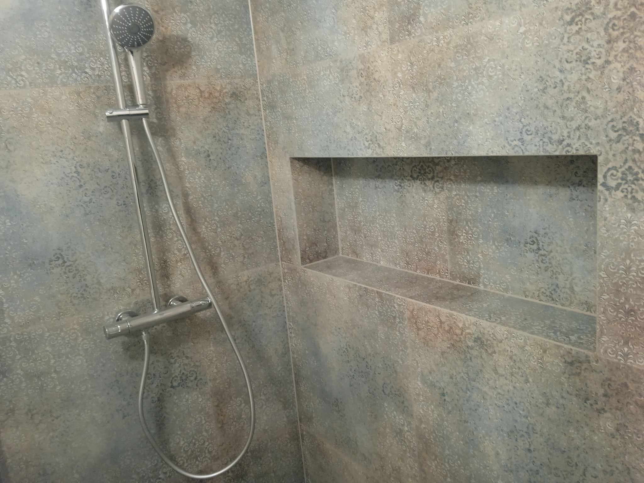 Kabina prysznicowa wyłożona płytkami z delikatnym, ornamentacyjnym wzorem, wyposażona w chromowaną baterię i deszczownicę, z wnęką w ścianie pełniącą funkcję półki.