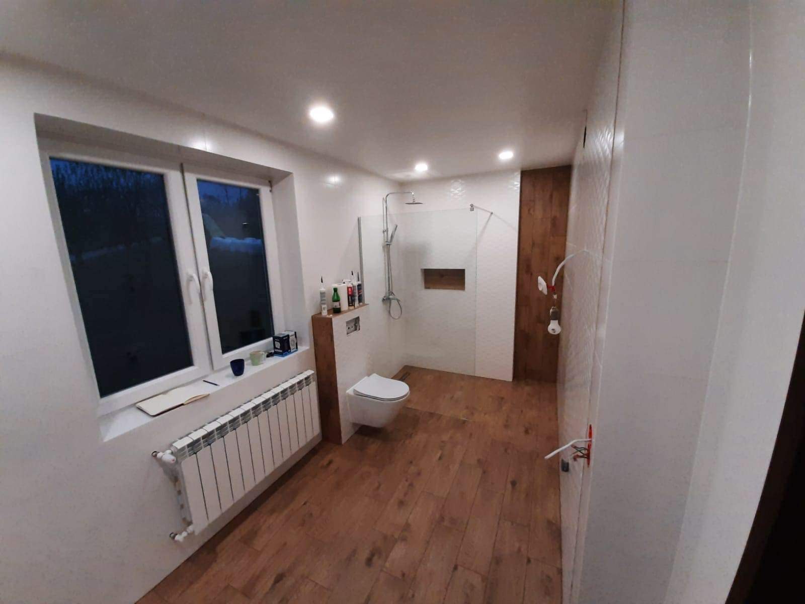 Nowoczesna łazienka z drewnopodobną podłogą, białą armaturą i kabiną prysznicową typu walk-in, widok na okno z grzejnikiem i akcesoriami łazienkowymi na półce