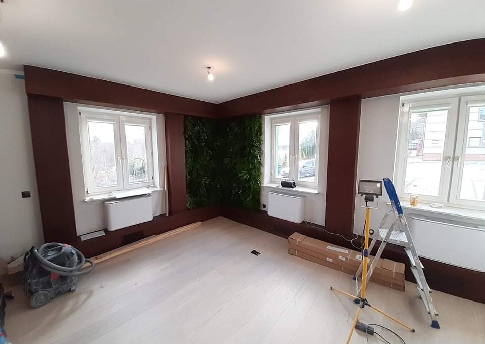 Pokój po częściowym remoncie z drewnianymi panelami, trzema oknami, ścianą z zieleni, odkurzaczem, drabiną i lampą budowlaną.