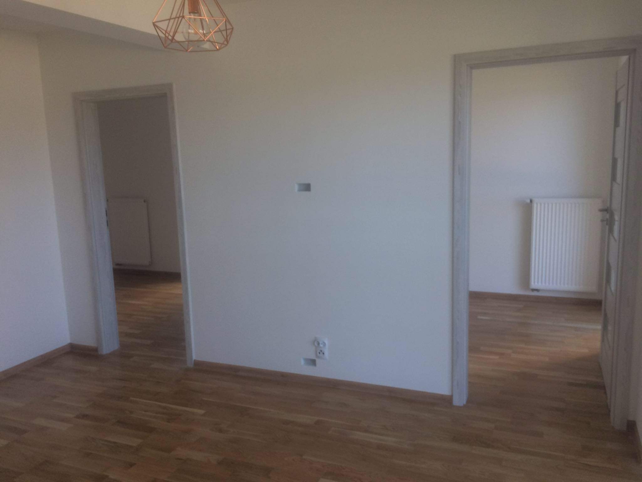 Pusty pokój po remoncie z parkietem, dwoma otworami drzwiowymi z szarymi ościeżnicami i wiszącą lampą w geometrycznym stylu.